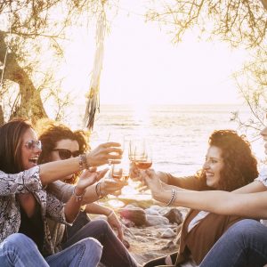 Group of middle age young adult women having fun together toasting and clinking with wine glasses at the beach during a golden sunset enjoying outdoor leisure activity or vacation